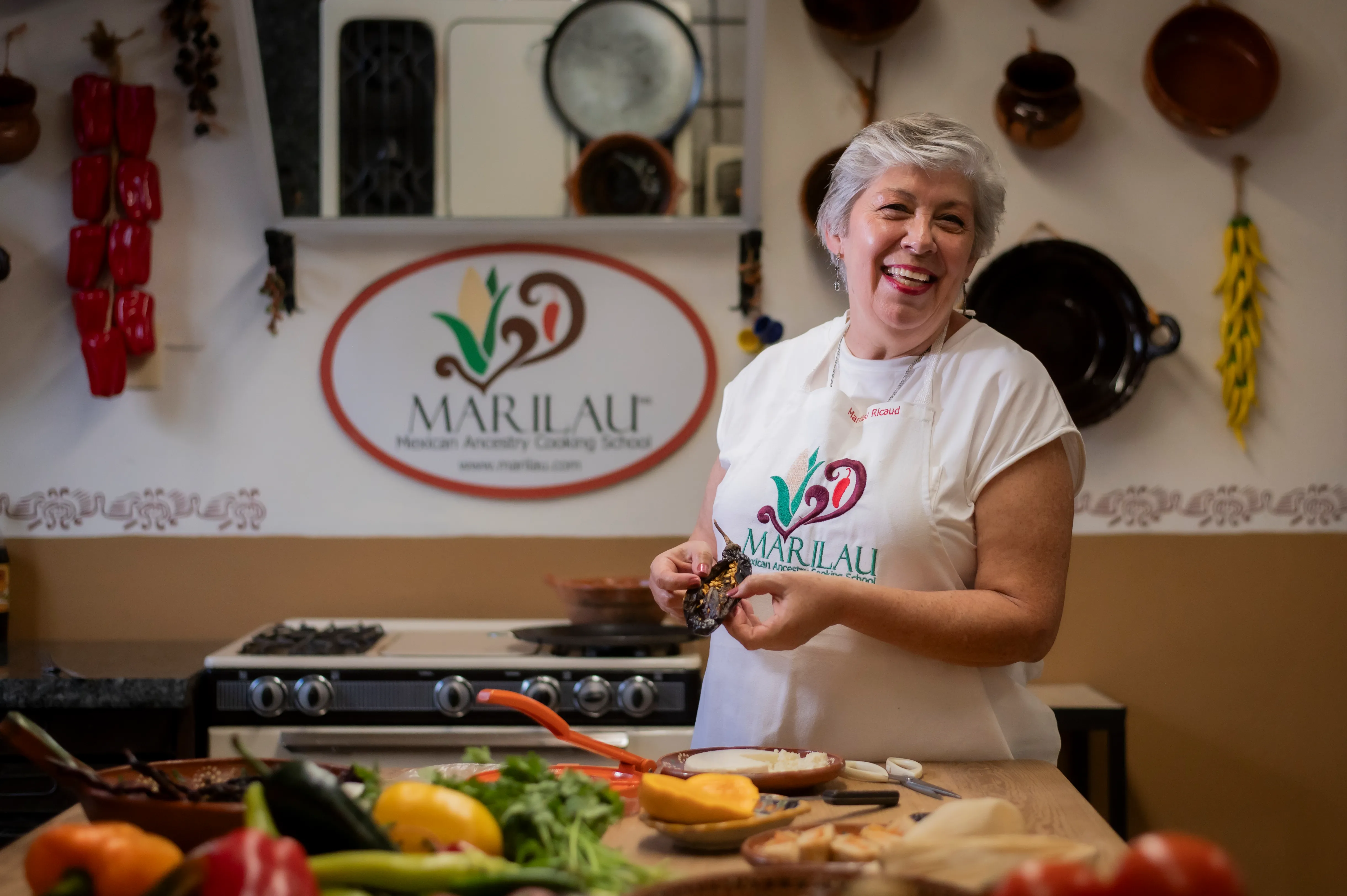 Marilau laughing in her kitchen, wearing the Marilau Mexican Ancestry Cooking School apron